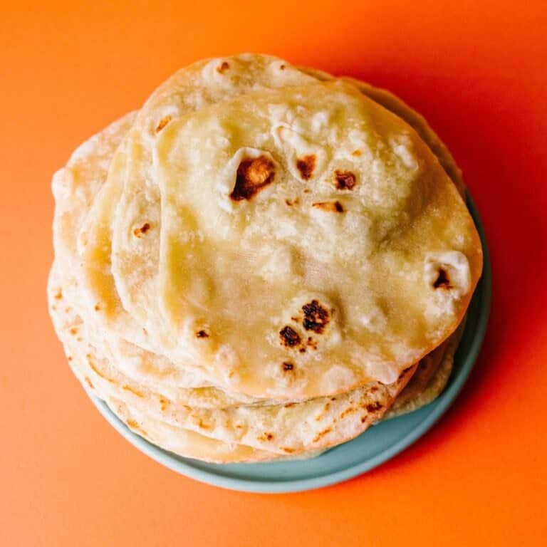Flour tortilla wrap recipe. Soft, homemade flour tortillas stacked on a wooden board. The tortillas have golden brown spots, a light and fluffy texture, and are wrapped in a cloth to keep warm.