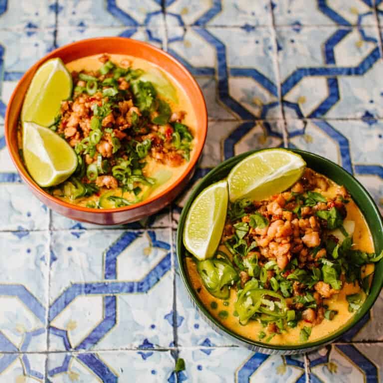 A bowl of spicy pork Thai coconut curry ramen, topped with crispy caramelised pork, fresh coriander, sliced red chillies, and spring onions, served with lime wedges on the side. The creamy coconut curry broth with ramen noodles and vibrant pak choi.